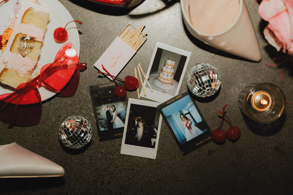 A photo shows a wedding detail flat lay including heart shaped glasses, polaroids, disco balls, and matches taken by a cake slice at a Long Branch NJ wedding held at The Wave venue 