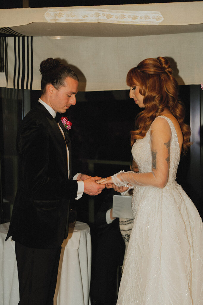A groom in a black tux exchanges rings with his bride during their wedding ceremony held at The Wave venue 