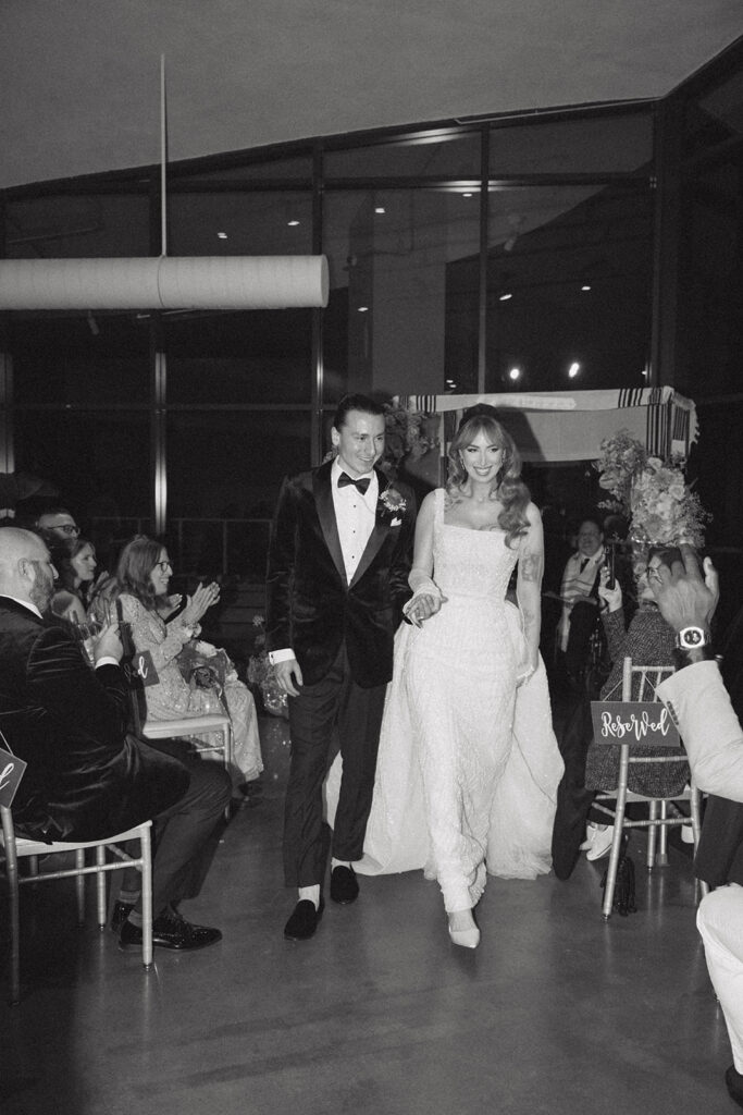 A black and white image shows newlyweds walking hand-in-hand down the aisle at their wedding in Long Branch NJ 