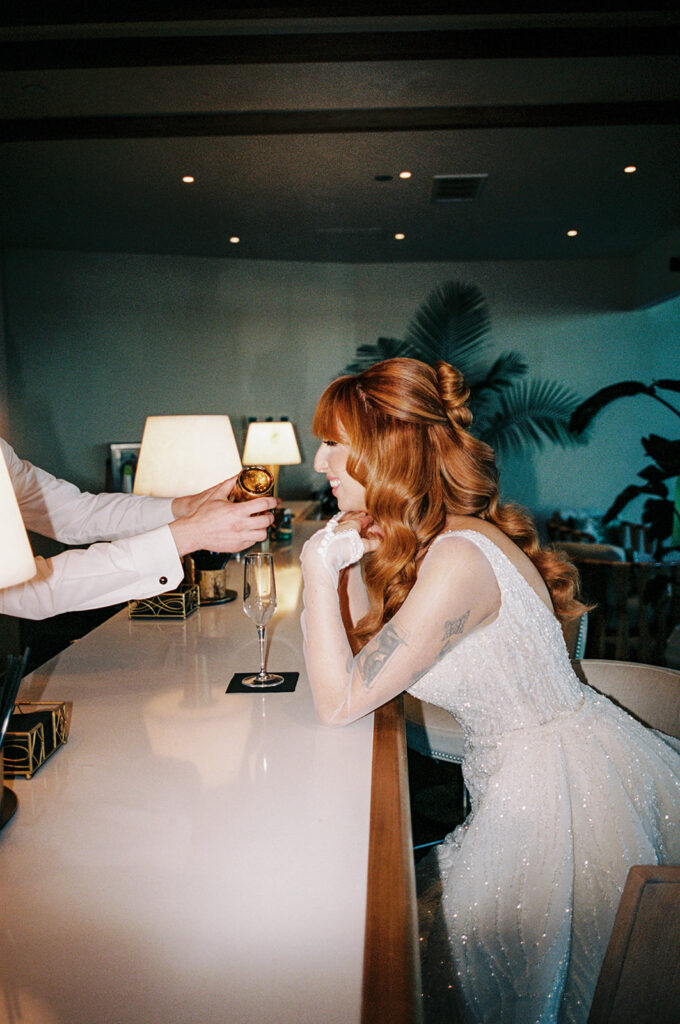 A bride in her gown rests her elbows on the bar at The Wave Resort as she awaits a glass of wine at her wedding 