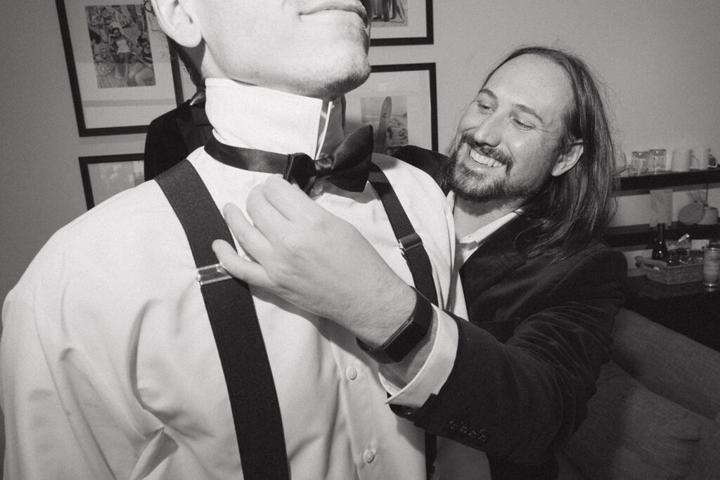 A black and white image shows a groomsman assisting the groom with his tie before his wave resort long branch wedding