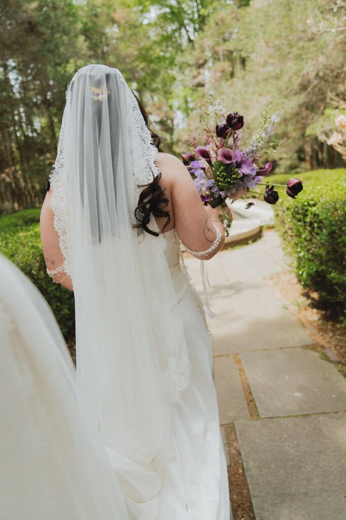 A bride in a long lace veil carries a purple bouquet walks toward her groom for first looks at their ridley creek wedding