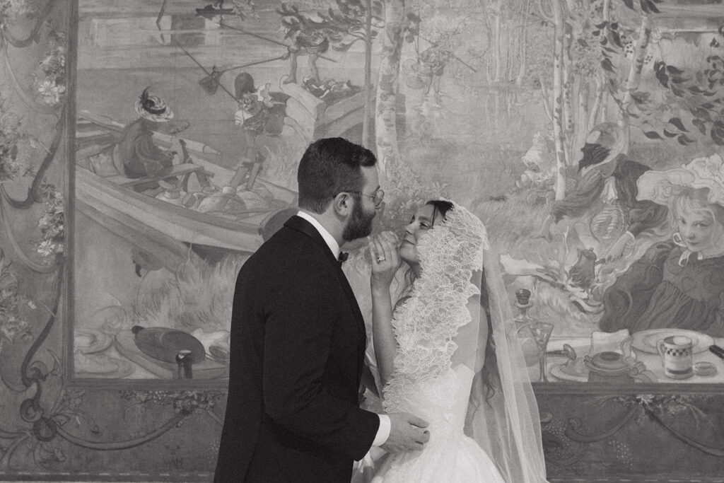 A black and white image depicts a bride and groom standing before a mural at their American Swedish Historical Museum wedding